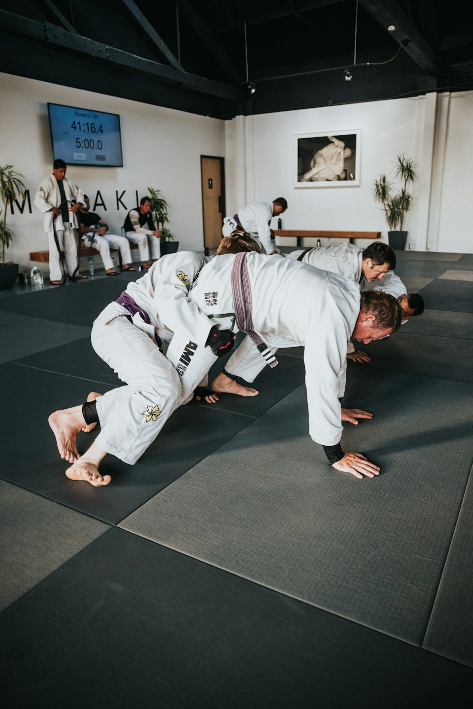 Hombres con gi blancos haciendo lagartijas de jiu-jitsu brasileño sobre tapetes grises.